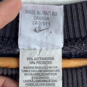 May include: A close-up of a label from a dark grey sweatshirt. The label states "MADE IN CANADA" and features the Nike logo. It details the fabric composition as 90% cotton and 10% polyester, plus washing instructions. The size is XXL/TTG.