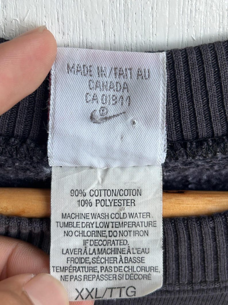 May include: A close-up of a label from a dark grey sweatshirt. The label states "MADE IN CANADA" and features the Nike logo. It details the fabric composition as 90% cotton and 10% polyester, plus washing instructions. The size is XXL/TTG.