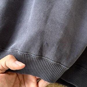 May include: Close-up of a dark grey sweatshirt. The ribbed hem is visible, along with the stitching. The fabric appears soft and slightly worn. The sweatshirt is a solid colour, with no visible graphics or patterns.