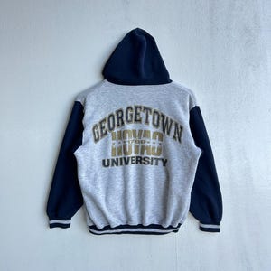May include: A gray hooded sweatshirt with navy blue sleeves and hood. The back of the sweatshirt features the words "GEORGETOWN UNIVERSITY" in gold lettering. The sweatshirt has a striped hem and cuffs.