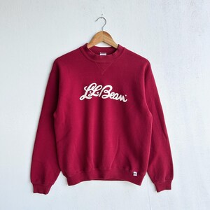 May include: A maroon crewneck sweatshirt with the white cursive text "L.L.Bean" across the chest. The sweatshirt is hanging on a wooden hanger against a white background. A small white tag is visible on the lower right sleeve.