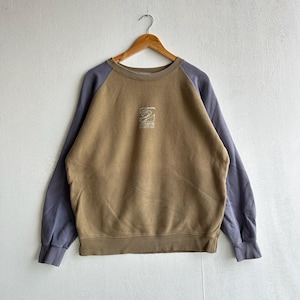 Könnte beinhalten: Ein zweifarbiges Sweatshirt in Braun und Blau. Der Körper des Sweatshirts ist braun, mit blauen Ärmeln. Eine weiße Grafik ist auf der Brust zentriert. Das Sweatshirt hängt an einem Holzbügel.