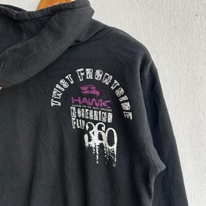 Peut inclure: Sweat &agrave; capuche noir avec cordon de serrage. Le dos du sweat &agrave; capuche porte l'inscription blanche et violette "TWIST FRONTSIDE HAWK NOSEGRIND FLIP 360". Le sweat &agrave; capuche est suspendu &agrave; un cintre en bois.