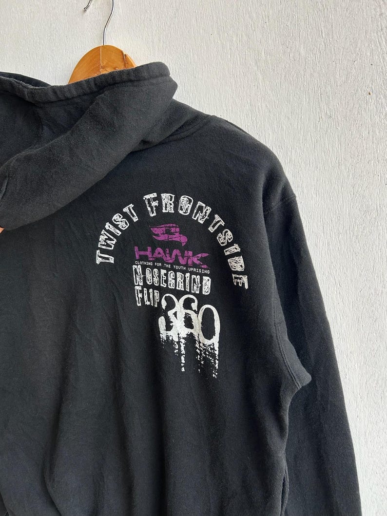 Peut inclure: Sweat &agrave; capuche noir avec cordon de serrage. Le dos du sweat &agrave; capuche porte l'inscription blanche et violette "TWIST FRONTSIDE HAWK NOSEGRIND FLIP 360". Le sweat &agrave; capuche est suspendu &agrave; un cintre en bois.