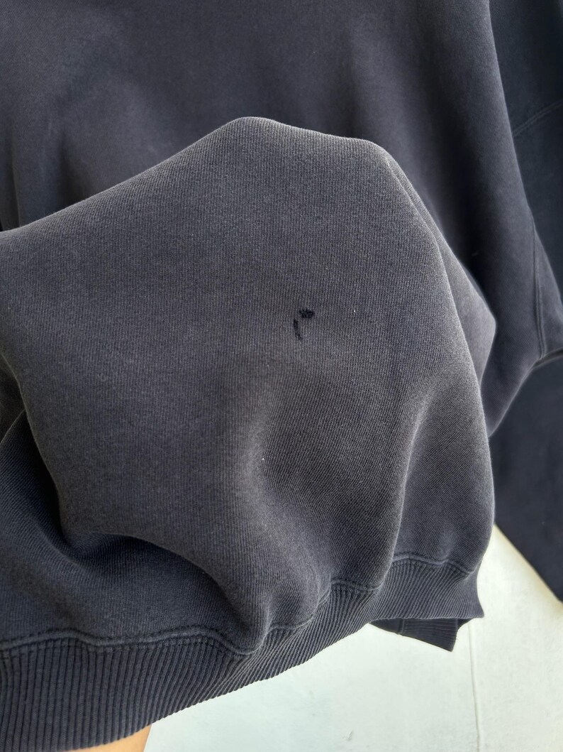 May include: Close-up of a dark navy blue sweatshirt. The fabric is slightly wrinkled, with a small hole visible. The bottom hem and cuffs are ribbed. The sweatshirt appears to be made of a soft, comfortable material.