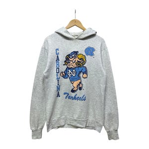 K&ouml;nnte beinhalten: Ein graues Kapuzensweatshirt mit einem Cartoon-Widder-Maskottchen in Blau und Gold, mit dem Text "UNC" und "Tarheels". Das Wort "Carolina" ist vertikal auf der linken Seite des Sweatshirts aufgedruckt.