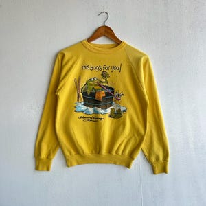 May include: Yellow sweatshirt with a graphic of a frog in a barrel with the text "this bug's for you!". The sweatshirt has long sleeves and a crew neck. The graphic also includes the text "Glenwood Springs Colorado".