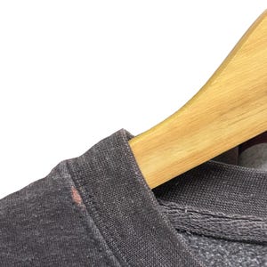 May include: Close-up of a dark gray sweatshirt with a crew neck. The fabric has a textured appearance. A wooden hanger is partially visible, supporting the garment. A small stain is visible on the shoulder.