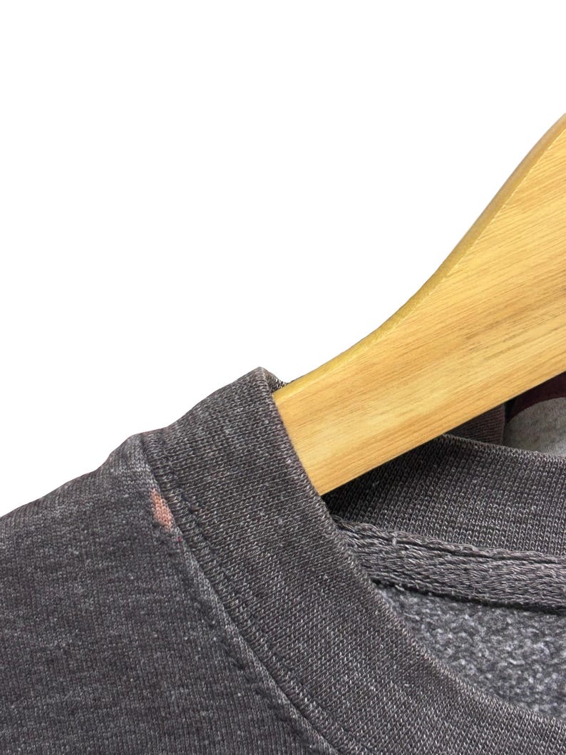 May include: Close-up of a dark gray sweatshirt with a crew neck. The fabric has a textured appearance. A wooden hanger is partially visible, supporting the garment. A small stain is visible on the shoulder.