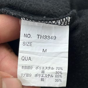 Peut inclure: Gros plan sur une &eacute;tiquette de v&ecirc;tement noire avec texte blanc. L'&eacute;tiquette indique "NO. TH3349" et "SIZE M". Le texte suppl&eacute;mentaire d&eacute;taille la composition du tissu, avec les pourcentages de polyester et de coton.