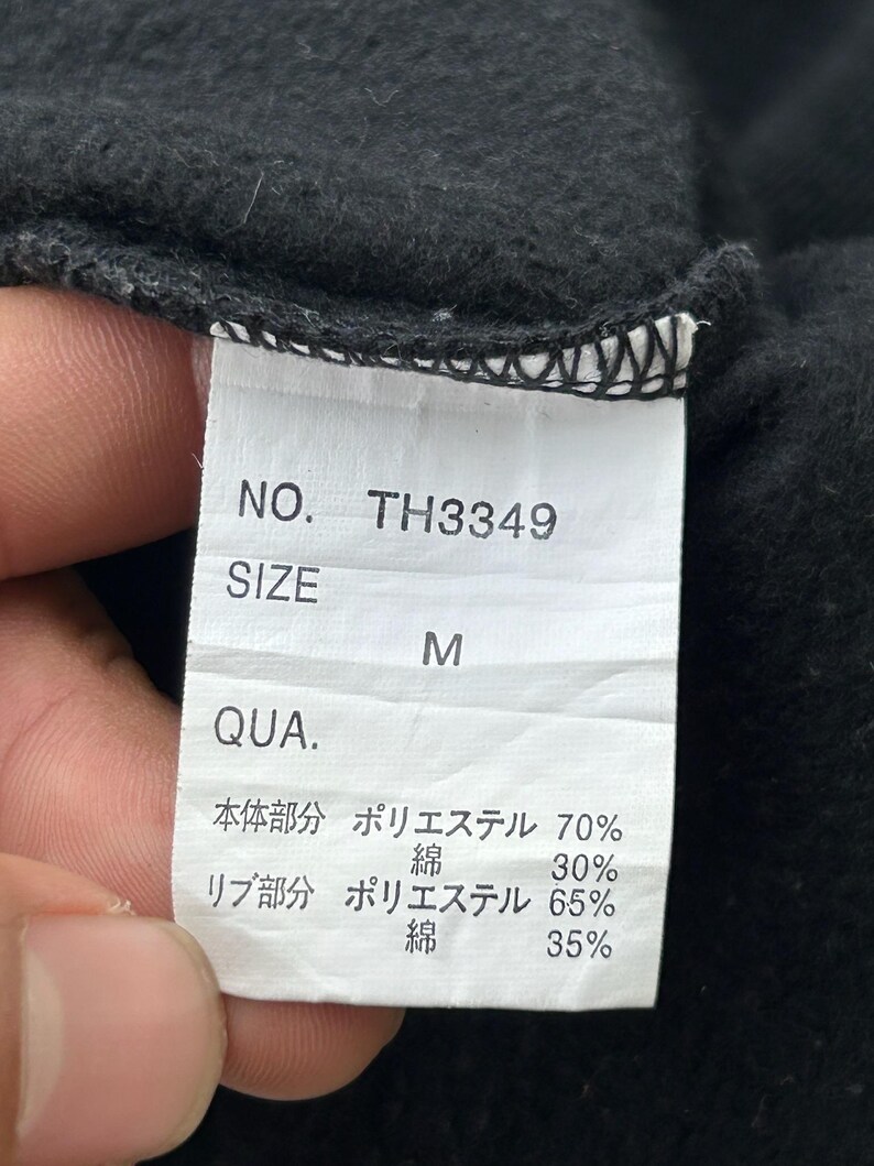 Peut inclure: Gros plan sur une &eacute;tiquette de v&ecirc;tement noire avec texte blanc. L'&eacute;tiquette indique "NO. TH3349" et "SIZE M". Le texte suppl&eacute;mentaire d&eacute;taille la composition du tissu, avec les pourcentages de polyester et de coton.