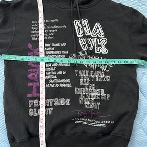 Peut inclure: Sweat &agrave; capuche noir avec texte blanc et violet "HAWK" et "Tony Hawk". Le sweat &agrave; capuche a une capuche &agrave; cordon et des manches longues. Le texte comprend des phrases comme "Frontside Blunt" et "Clothing for the Youth Uprising".