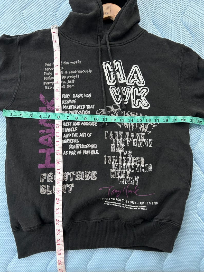 Peut inclure: Sweat &agrave; capuche noir avec texte blanc et violet "HAWK" et "Tony Hawk". Le sweat &agrave; capuche a une capuche &agrave; cordon et des manches longues. Le texte comprend des phrases comme "Frontside Blunt" et "Clothing for the Youth Uprising".