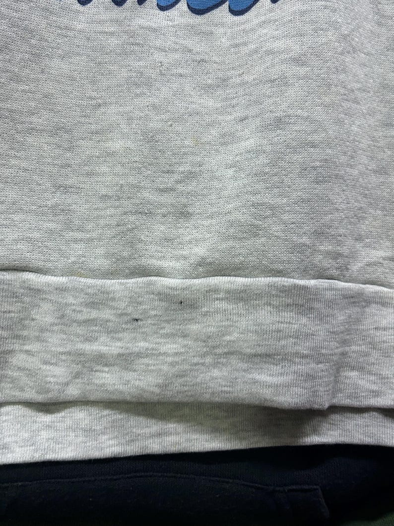 K&ouml;nnte beinhalten: Ein graues Sweatshirt mit blauem Text oben. Der untere Teil des Sweatshirts ist schwarz. Das Sweatshirt hat einen gerippten Saum. Der Stoff scheint ein weiches, bequemes Material zu sein.