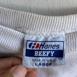 May include: Close-up of a white crew neck shirt with a tag that reads "Hanes BEEFY MADE IN U.S.A. LARGE". The tag is blue and white with the Hanes logo.