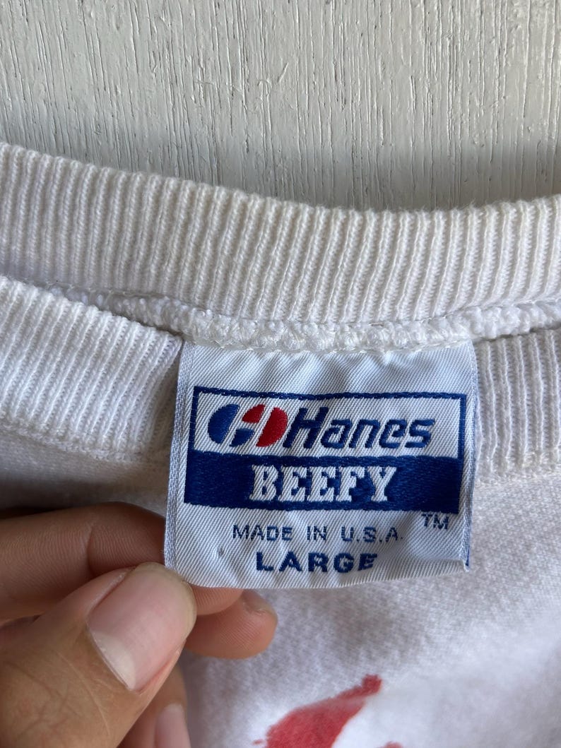 May include: Close-up of a white crew neck shirt with a tag that reads "Hanes BEEFY MADE IN U.S.A. LARGE". The tag is blue and white with the Hanes logo.