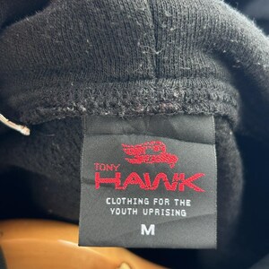 Peut inclure: Gros plan sur une &eacute;tiquette de sweat &agrave; capuche noire avec le texte "TONY HAWK" en rouge et "CLOTHING FOR THE YOUTH UPRISING" en dessous. L'&eacute;tiquette porte &eacute;galement la lettre "M" pour medium.