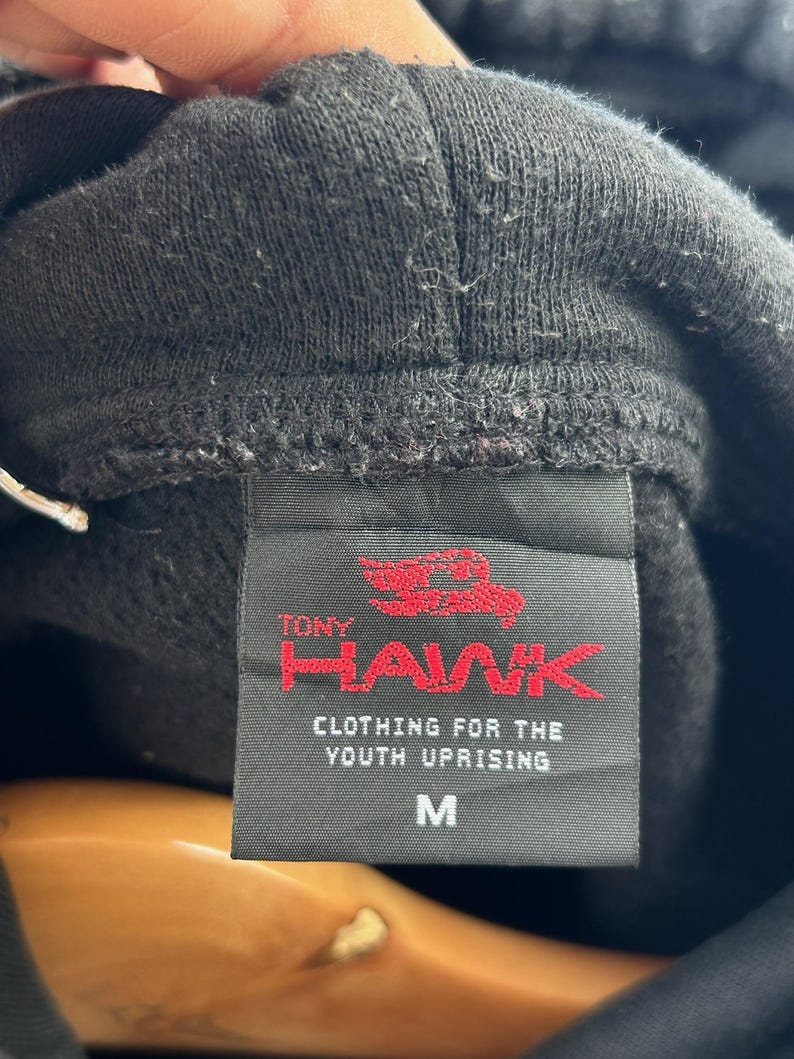 Peut inclure: Gros plan sur une &eacute;tiquette de sweat &agrave; capuche noire avec le texte "TONY HAWK" en rouge et "CLOTHING FOR THE YOUTH UPRISING" en dessous. L'&eacute;tiquette porte &eacute;galement la lettre "M" pour medium.