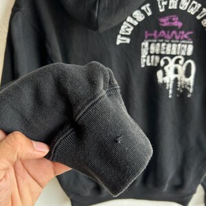 Peut inclure: Sweat &agrave; capuche noir avec texte graphique blanc et violet au dos. Le texte comprend les mots "Twist Front" et "Hawk". Le poignet de la manche est visible au premier plan.