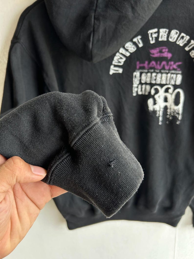 Peut inclure: Sweat &agrave; capuche noir avec texte graphique blanc et violet au dos. Le texte comprend les mots "Twist Front" et "Hawk". Le poignet de la manche est visible au premier plan.