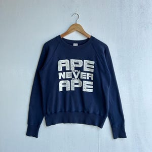 Könnte beinhalten: Marineblaues Sweatshirt mit dem weißen Text "APE SHALL NEVER KILL APE". Das Sweatshirt mit Rundhalsausschnitt hat lange Ärmel. Das Kleidungsstück hängt an einem Holzbügel.
