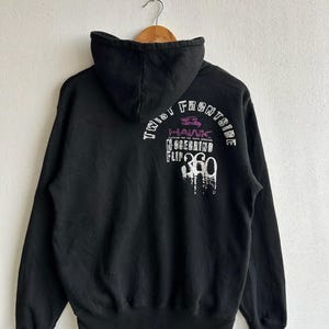 Peut inclure: Sweat &agrave; capuche noir avec cordon de serrage, avec texte blanc et violet au dos. Le texte comprend "TWIST FRONTSIDE", "HAWK", "NOSEGRIND" et "FLIP 360". Le sweat &agrave; capuche est suspendu &agrave; un cintre en bois.