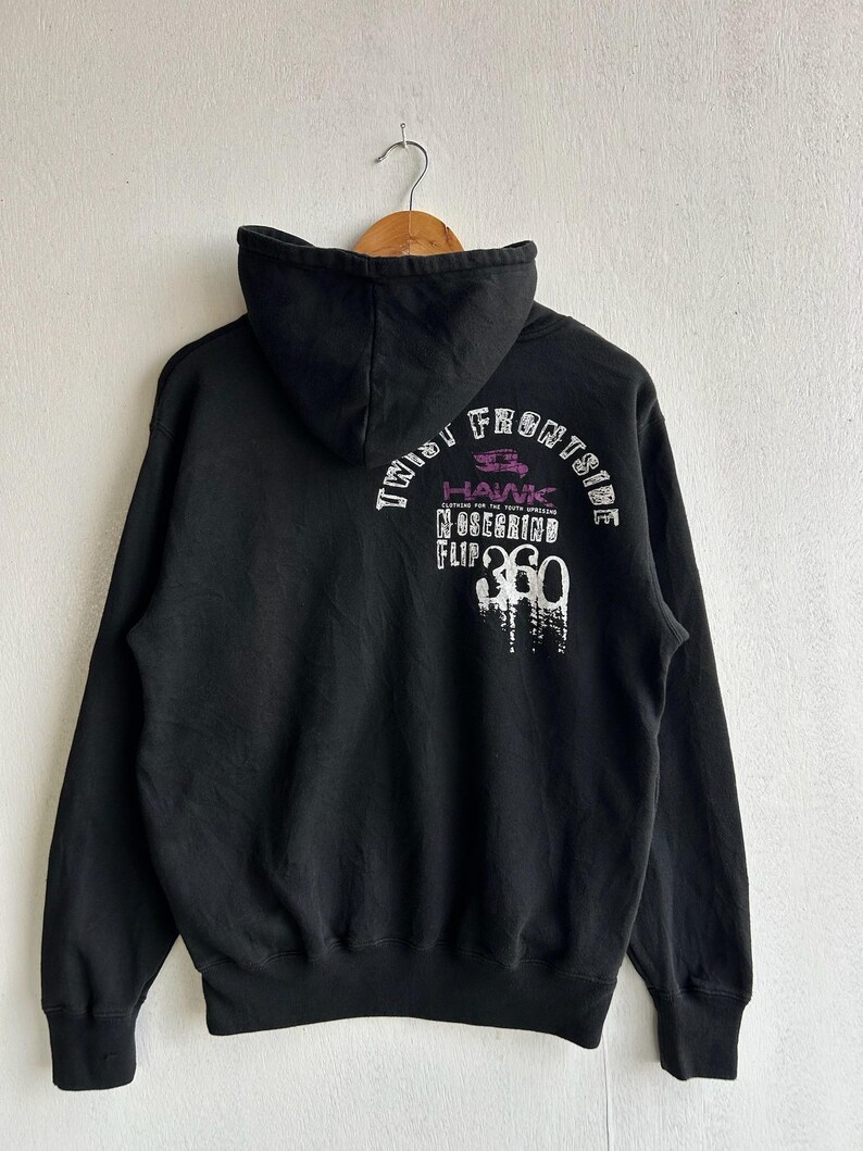 Peut inclure: Sweat &agrave; capuche noir avec cordon de serrage, avec texte blanc et violet au dos. Le texte comprend "TWIST FRONTSIDE", "HAWK", "NOSEGRIND" et "FLIP 360". Le sweat &agrave; capuche est suspendu &agrave; un cintre en bois.