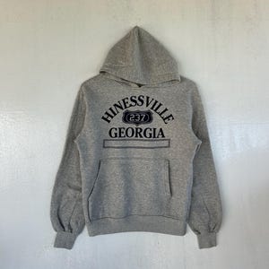 Puede incluir: Sudadera con capucha gris con bolsillo delantero y el texto "Hinesville Georgia 237" en azul marino. La sudadera tiene mangas largas y una capucha con cordón.
