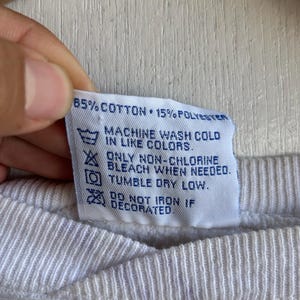 May include: Close-up of a white fabric tag with care instructions. The tag reads "85% COTTON 15% POLYESTER" and includes washing symbols for machine wash cold, non-chlorine bleach, and tumble dry low.