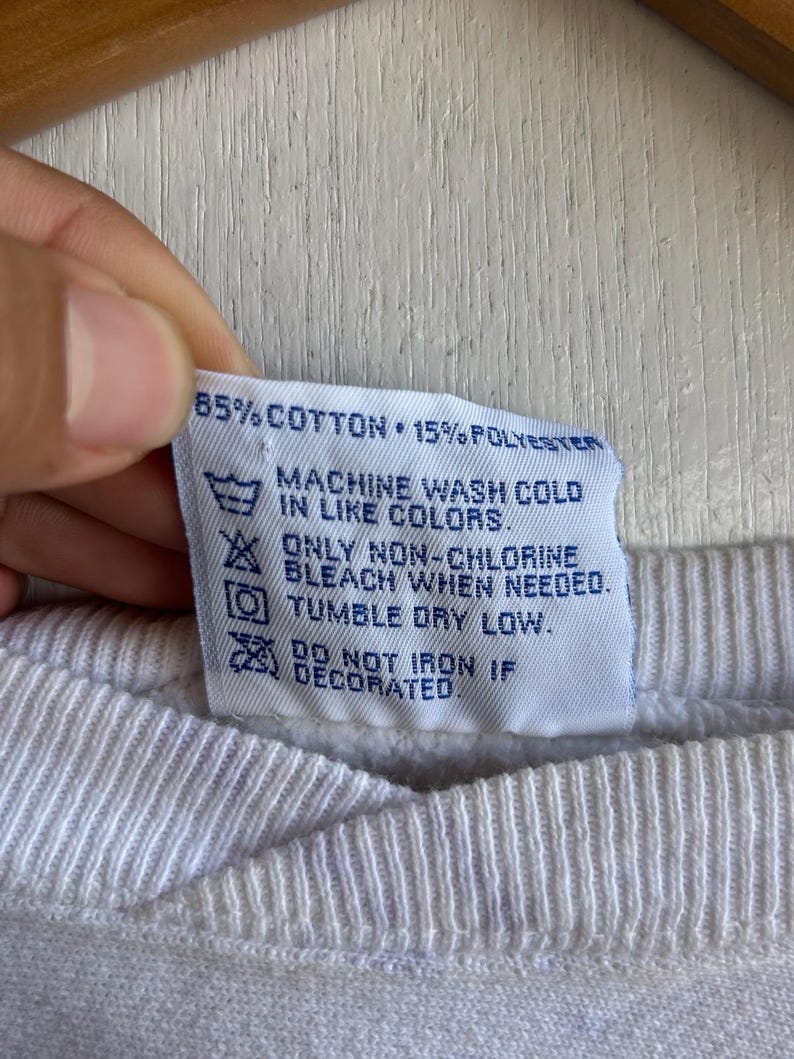 May include: Close-up of a white fabric tag with care instructions. The tag reads "85% COTTON 15% POLYESTER" and includes washing symbols for machine wash cold, non-chlorine bleach, and tumble dry low.