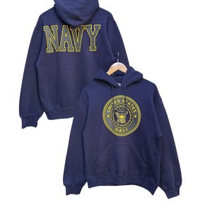 Könnte beinhalten: Marineblauer Hoodie mit Fronttasche und Kapuze. Auf der Vorderseite befindet sich ein gelb-schwarzes Emblem mit den Worten "UNITED STATES NAVY". Auf der Rückseite des Hoodies steht das Wort "NAVY" in großen gelben Buchstaben.