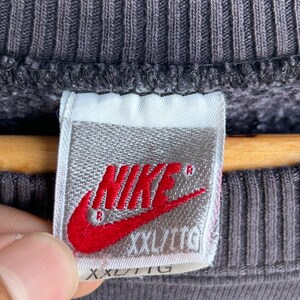 May include: A close-up shot of a grey sweatshirt with a ribbed texture. A white label with the red Nike logo and the text "XXL/TTG" is visible. The label is attached to the sweatshirt.
