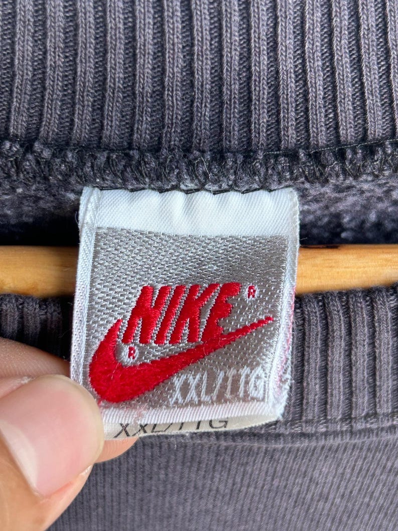 May include: A close-up shot of a grey sweatshirt with a ribbed texture. A white label with the red Nike logo and the text "XXL/TTG" is visible. The label is attached to the sweatshirt.