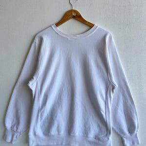May include: A white crew neck sweatshirt with long sleeves, hanging on a wooden hanger. The sweatshirt has ribbed cuffs and a ribbed hem. The fabric appears to be a soft, comfortable material.