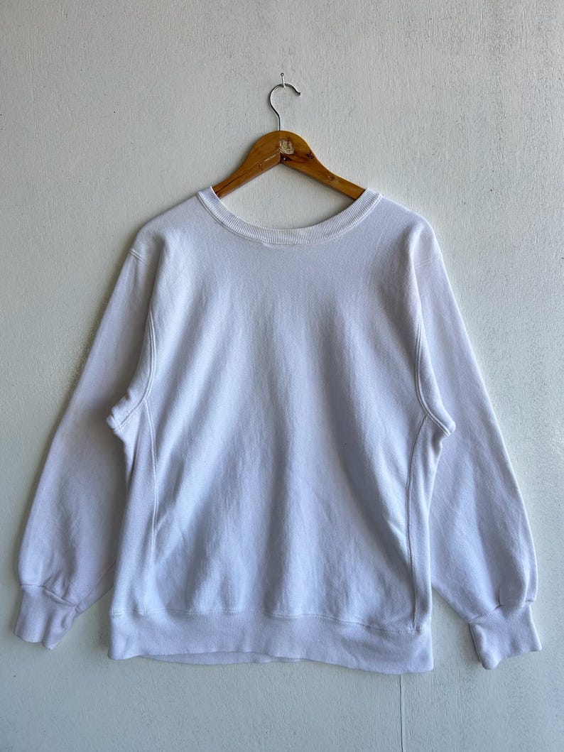 May include: A white crew neck sweatshirt with long sleeves, hanging on a wooden hanger. The sweatshirt has ribbed cuffs and a ribbed hem. The fabric appears to be a soft, comfortable material.