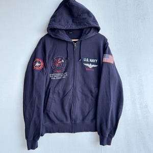 May include: Navy blue zip-up hoodie with a hood. Features the text "U.S. NAVY" and an American flag patch on the right sleeve. Includes patches and text on the left chest and sleeve. A casual outerwear garment.