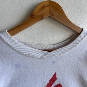 May include: White crew neck sweatshirt with a red graphic design. The sweatshirt is hanging on a wooden hanger against a white background. The design is a stylized, abstract shape.