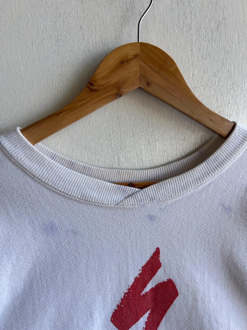 May include: White crew neck sweatshirt with a red graphic design. The sweatshirt is hanging on a wooden hanger against a white background. The design is a stylized, abstract shape.