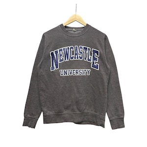 May include: A charcoal gray sweatshirt with the words "NEWCASTLE UNIVERSITY" in a blue and white arched design across the chest. The sweatshirt is hanging on a wooden hanger against a white background.
