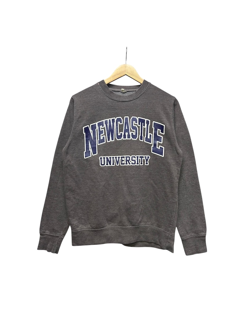 May include: A charcoal gray sweatshirt with the words "NEWCASTLE UNIVERSITY" in a blue and white arched design across the chest. The sweatshirt is hanging on a wooden hanger against a white background.