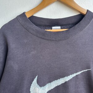 May include: A dark grey sweatshirt featuring a light blue embroidered Nike swoosh logo. The crew neck sweatshirt is displayed on a wooden hanger. The fabric appears to be a soft, worn material.