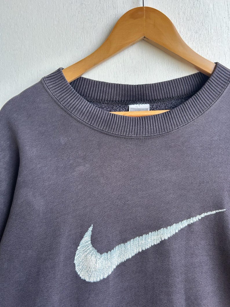 May include: A dark grey sweatshirt featuring a light blue embroidered Nike swoosh logo. The crew neck sweatshirt is displayed on a wooden hanger. The fabric appears to be a soft, worn material.