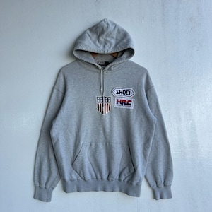 Puede incluir: Una sudadera con capucha gris con bolsillo de canguro y cordón. La sudadera presenta parches con el texto "SHOEI" y "HRC" junto con un diseño de escudo con rayas rojas y blancas y estrellas azules.