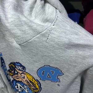 K&ouml;nnte beinhalten: Ein hellgrauer Kapuzenpullover mit einem Cartoon-Maskottchen und den Buchstaben "UNC" in Blau und Gelb. Der Pullover hat auch ein hellblaues "NC"-Logo. Der &Auml;rmel weist Gebrauchsspuren auf.