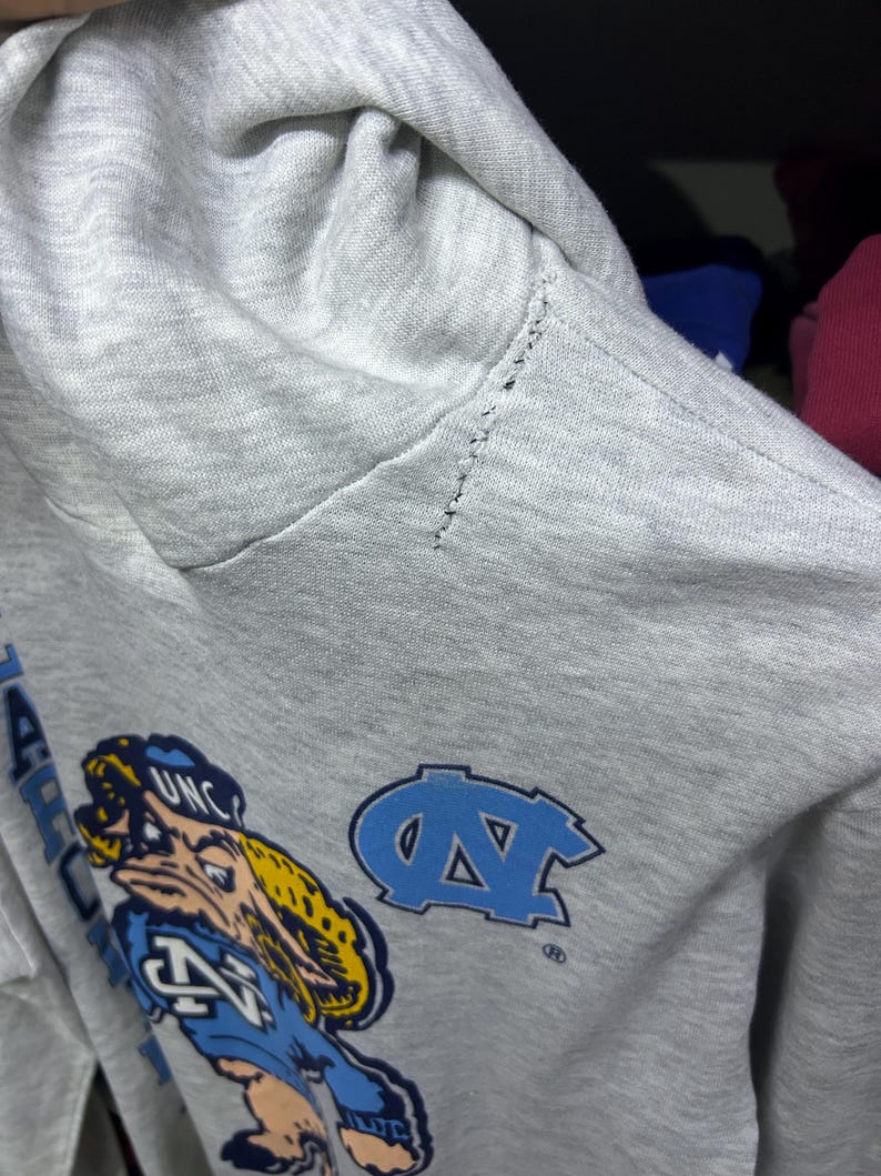 K&ouml;nnte beinhalten: Ein hellgrauer Kapuzenpullover mit einem Cartoon-Maskottchen und den Buchstaben "UNC" in Blau und Gelb. Der Pullover hat auch ein hellblaues "NC"-Logo. Der &Auml;rmel weist Gebrauchsspuren auf.