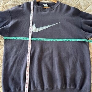 May include: A dark grey Nike sweatshirt featuring a large white swoosh logo. The sweatshirt is laid flat, with a measuring tape showing a width of approximately 68.5 cm. The garment is on a wooden hanger.