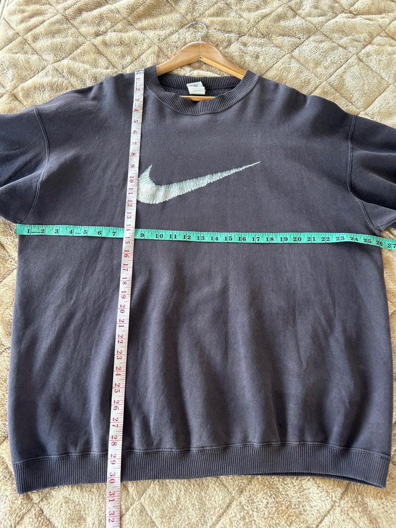 May include: A dark grey Nike sweatshirt featuring a large white swoosh logo. The sweatshirt is laid flat, with a measuring tape showing a width of approximately 68.5 cm. The garment is on a wooden hanger.