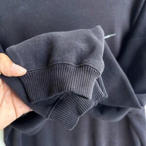 May include: Close-up of a dark grey sweatshirt sleeve. The ribbed cuff is visible, along with a small white logo. The fabric appears soft and slightly worn. The sweatshirt is a casual apparel item.