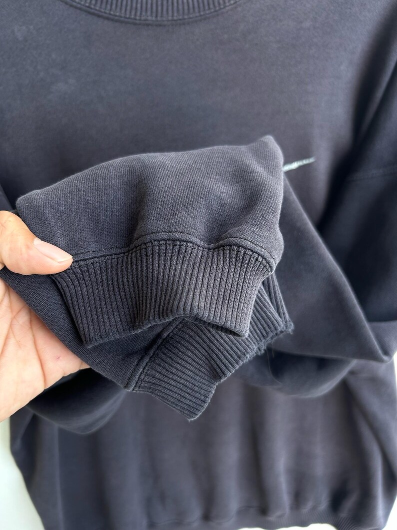 May include: Close-up of a dark grey sweatshirt sleeve. The ribbed cuff is visible, along with a small white logo. The fabric appears soft and slightly worn. The sweatshirt is a casual apparel item.