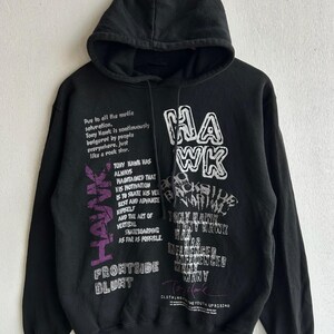 Peut inclure: Sweat &agrave; capuche noir avec cordon de serrage et manches longues. Le devant affiche le mot "HAWK" en blanc et violet, ainsi que du texte sur Tony Hawk et le skateboard. Le sweat &agrave; capuche est en mati&egrave;re douce.